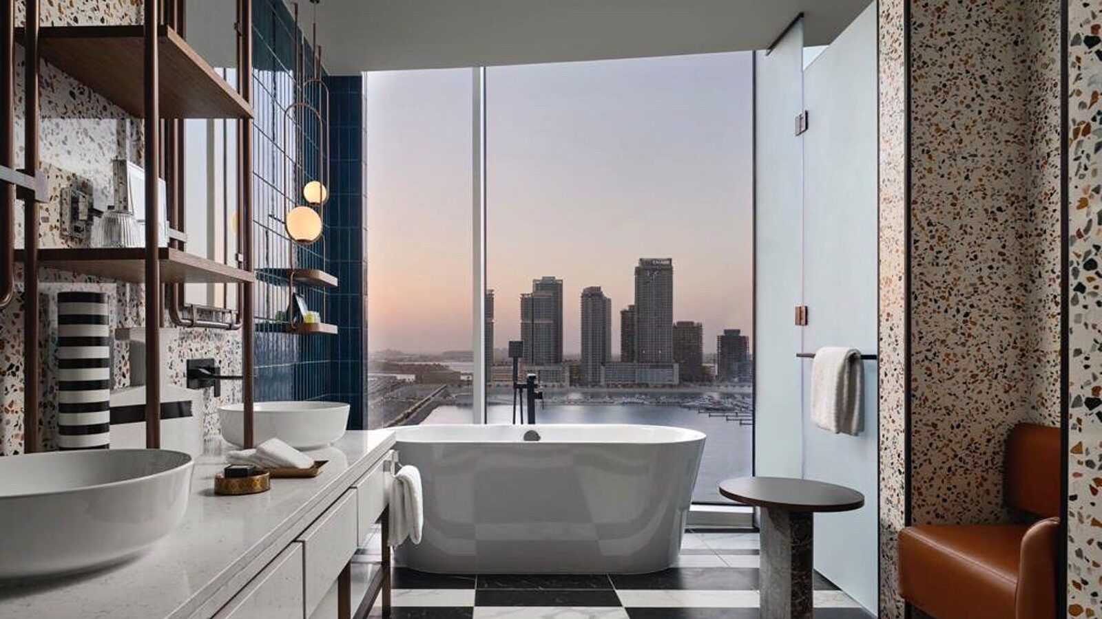 Freestanding bath in a terrazzo and tiled hotel bathroom with a city view at dusk